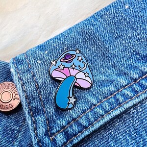 May include: A blue and pink enamel pin of a mushroom with a planet and stars on top. The pin is attached to a denim jacket.