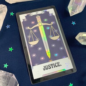 May include: A holographic tarot card with a purple background and a rainbow sword and scales. The card is labeled "XI JUSTICE."