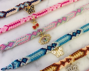 May include: Six colorful friendship bracelets with different charms, including a butterfly, a cherry, an evil eye, a butterfly with a crescent moon, a flower, and a cactus.