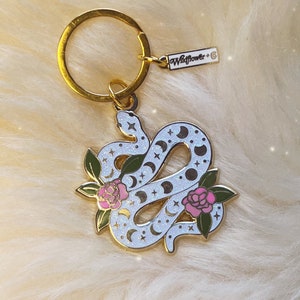 Może przedstawiać: Brelok do kluczy w kształcie złotego węża z białym emaliowanym wykończeniem i wzorem księżyca i gwiazd. Wąż jest zwinięty wokół dwóch różowych kwiatów z zielonymi liśćmi. Brelok ma złoty pierścień i metkę z napisem "Wildflower."