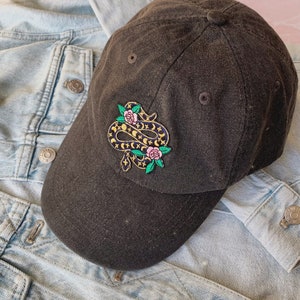 May include: A black baseball cap with a dark blue snake with pink flowers and gold stars embroidered on the front.