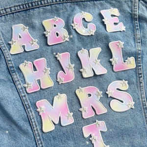 May include: A denim jacket with colorful letters A, B, C, E, H, J, K, L, M, R, S, and T. Each letter has a white outline and is decorated with silver glitter stars.