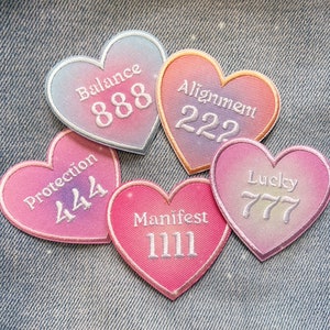 May include: Five pink and blue heart-shaped patches with white embroidery. The patches feature the words "Balance 888", "Alignment 222", "Protection 444", "Lucky 777", and "Manifest 1111".