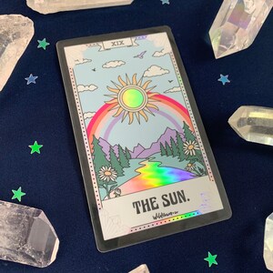Puede incluir: Una carta de tarot hologr&aacute;fica con un arco&iacute;ris y un sol en un cielo azul. La carta est&aacute; etiquetada como "XIX. EL SOL." y "Wildflower Co."