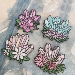 May include: Four embroidered iron-on patches featuring clusters of colourful crystals with floral accents. The crystals are in shades of pink, blue, and white, and the flowers are in shades of pink and white.