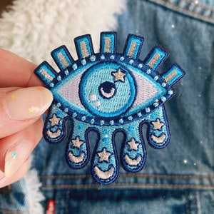 May include: A blue and white embroidered eye patch with a dripping design and stars. The eye patch is a popular symbol of protection and good luck.