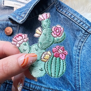 Peut inclure: Un patch brodé en forme de cactus vert avec des fleurs roses et des accents jaunes. Le patch est fixé sur une veste en jean bleue.