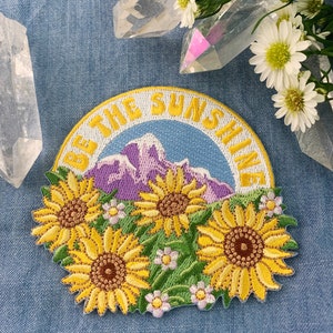 May include: Embroidered iron-on patch with a yellow, white, and green design of sunflowers and a mountain range. The text "Be the Sunshine" is in a white circle around the design.