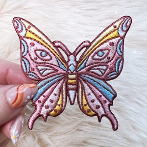 May include: A pink and blue embroidered butterfly patch with yellow accents. The butterfly has intricate details and a whimsical design.