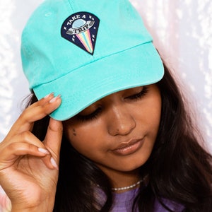 May include: A light turquoise baseball cap with a curved brim. The cap features a patch with a UFO design, the words "TAKE A TRIP", and a rainbow. The cap is made of a soft material.