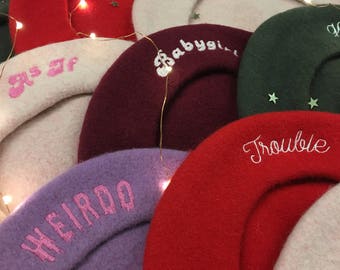May include: A stack of colorful berets with embroidered text, including "Babygirl", "Trouble", "Weirdo", and "as if".