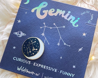 May include: A gold and black enamel pin featuring the Gemini constellation with a crescent moon. The pin is on a blue background with white stars and the text "Gemini" in a holographic font. The text "May 21 - June 20" is also on the background. The text "Curious. Expressive. Funny" is printed below the pin. The text "Wildflower+co" and "wildflower.co" are printed below the text.