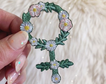 Könnte beinhalten: Grüner und weißer gestickter Aufnäher des weiblichen Geschlechtszeichens mit einem Kranz aus Blumen um die Oberseite.