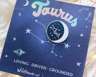 May include: A circular enamel pin featuring a dark blue crescent moon with a gold Taurus constellation design, mounted on a dark blue card printed with the word Taurus, the dates April 20-May 20, and the text LOVING DRIVEN GROUNDED.