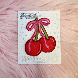 May include: An iron-on patch featuring a pair of red cherries with green stems and a pink bow. The patch is on a white card with the words "Iron-On Patch" and "Wildflower + Co." printed on it.