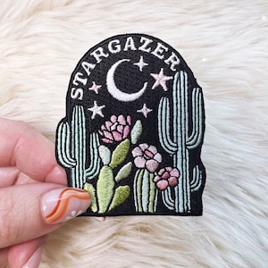 Peut inclure: Patch brodé avec un fond noir représentant une lune croissante, des étoiles, des cactus et des fleurs. Le texte "STARGAZER" est brodé en blanc autour du haut du patch.