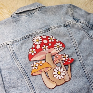 May include: A light blue denim jacket with a colorful mushroom patch. The patch features red, orange, and tan mushrooms with white daisy accents. The jacket has a classic collar, button closure, and pockets.
