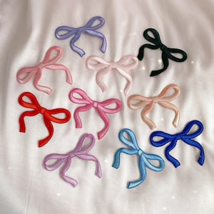 May include: Assortment of ten colorful fabric bows in various shades of pink, blue, red, and black. The bows are arranged on a white fabric background.
