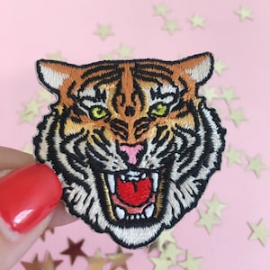 May include: Iron-on patch featuring an embroidered tiger's face. The tiger has orange, black and white fur and a fierce expression with a wide open mouth and sharp teeth.