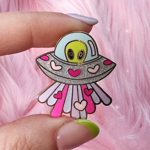 May include: A silver and blue enamel pin of a cartoon alien in a spaceship with pink hearts and glitter. The spaceship has a light blue dome and the alien has green skin and black eyes.