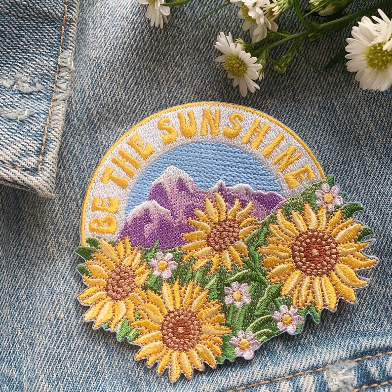 May include: Embroidered patch with a yellow, blue, and purple mountain scene and the text "Be the Sunshine". The patch features three yellow sunflowers with brown centers.