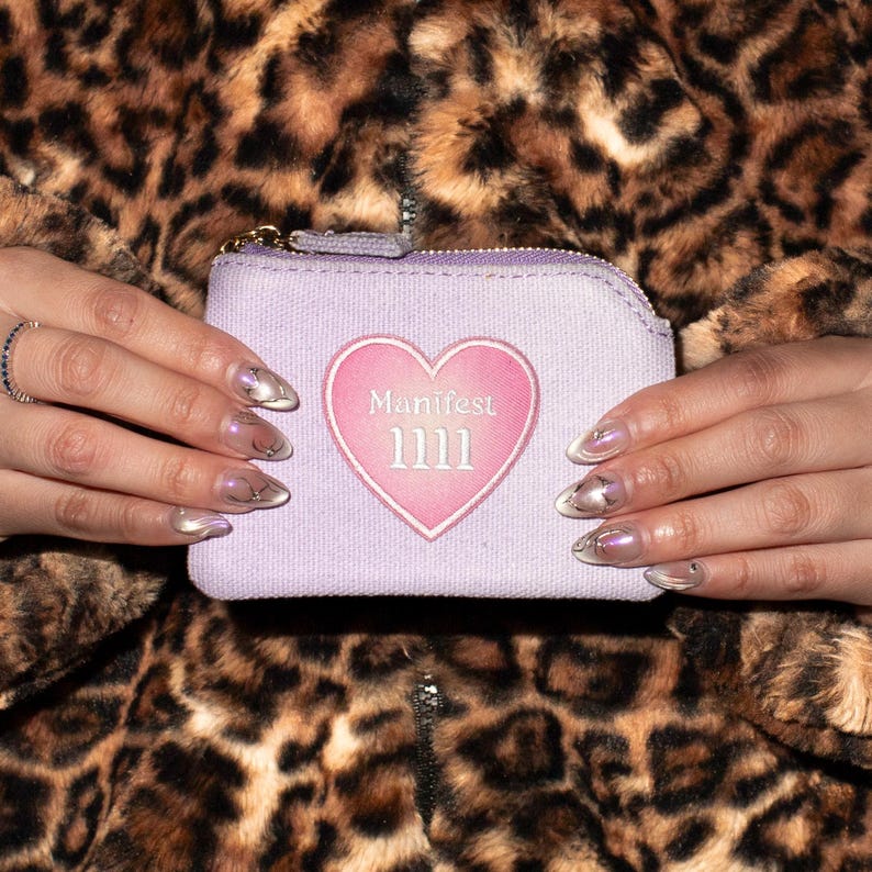 May include: A light purple zippered pouch with a pink heart design that reads "Manifest 1111". The pouch is held against a leopard print background. The pouch has a gold zipper and a small loop.
