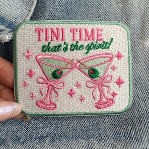 Peut inclure: Écusson brodé avec les mots "TINI TIME that's the spirit!" en rose et vert. L'écusson représente deux verres à martini avec des olives, des nœuds roses et des étoiles roses sur un fond blanc avec une bordure verte.