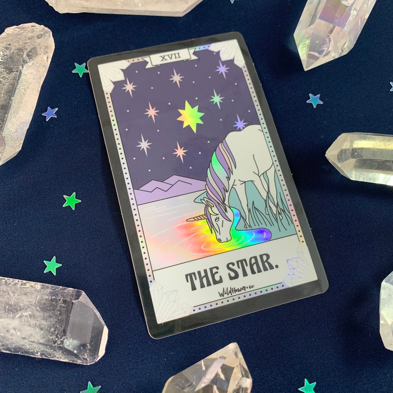 May include: A holographic tarot card with a silver border. The card is the Star card, number XVII. The card depicts a unicorn drinking from a pool of water with a rainbow reflection. The background is a dark purple sky with stars.