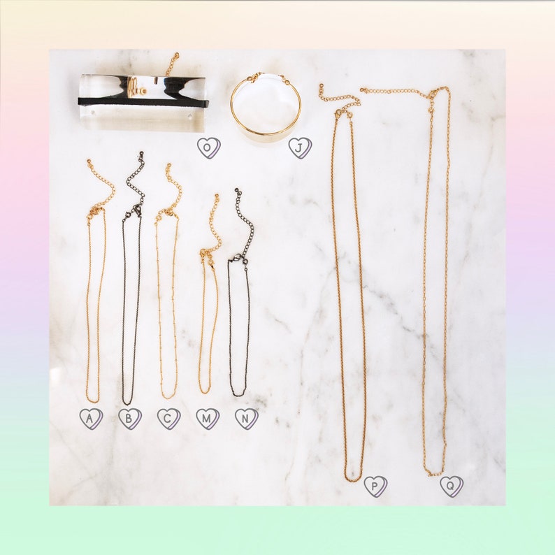 May include: A set of nine gold and black chain necklaces on a white marble background. The necklaces are labeled with letters A through Q.