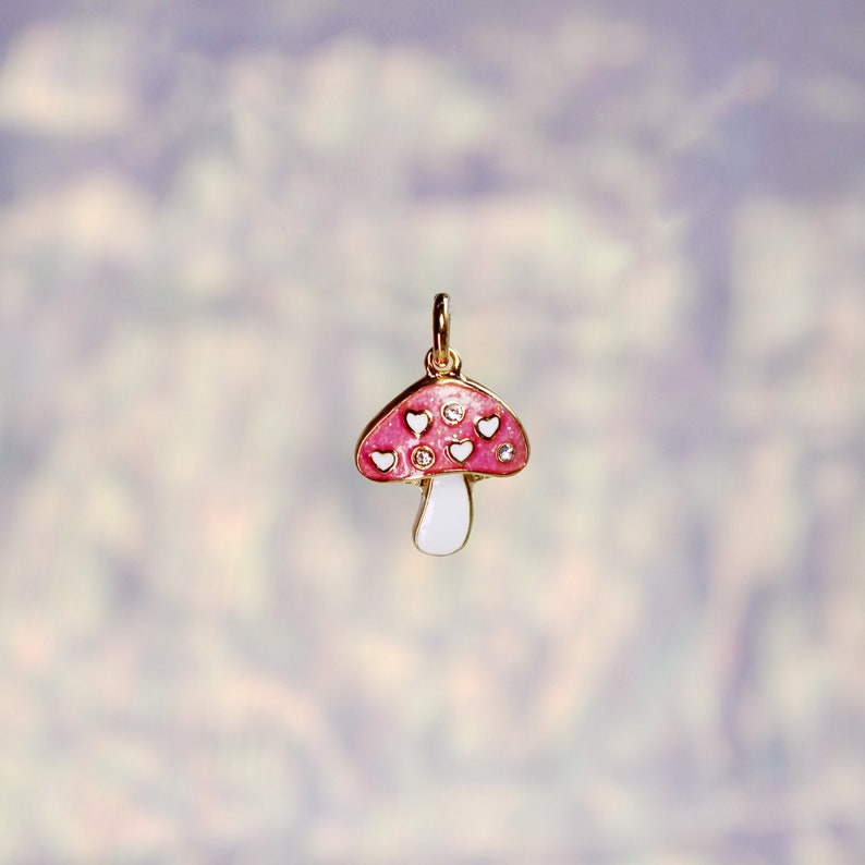 Pode incluir: Um amuleto de cogumelo de esmalte rosa e branco com detalhes dourados e cora&ccedil;&otilde;es brancos. O amuleto tem um caule branco e &eacute; perfeito para adicionar um toque caprichoso &agrave;s joias.
