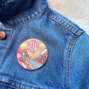 Peut inclure: Un &eacute;cusson rond brod&eacute; avec les mots "Stay Wild" en rose, repr&eacute;sentant un paysage d&eacute;sertique avec des cactus, des montagnes et une rivi&egrave;re. L'&eacute;cusson est cousu sur une veste en jean bleue.