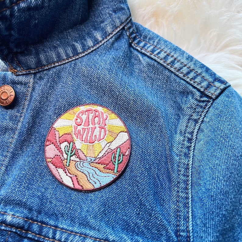 Peut inclure: Un &eacute;cusson rond brod&eacute; avec les mots "Stay Wild" en rose, repr&eacute;sentant un paysage d&eacute;sertique avec des cactus, des montagnes et une rivi&egrave;re. L'&eacute;cusson est cousu sur une veste en jean bleue.