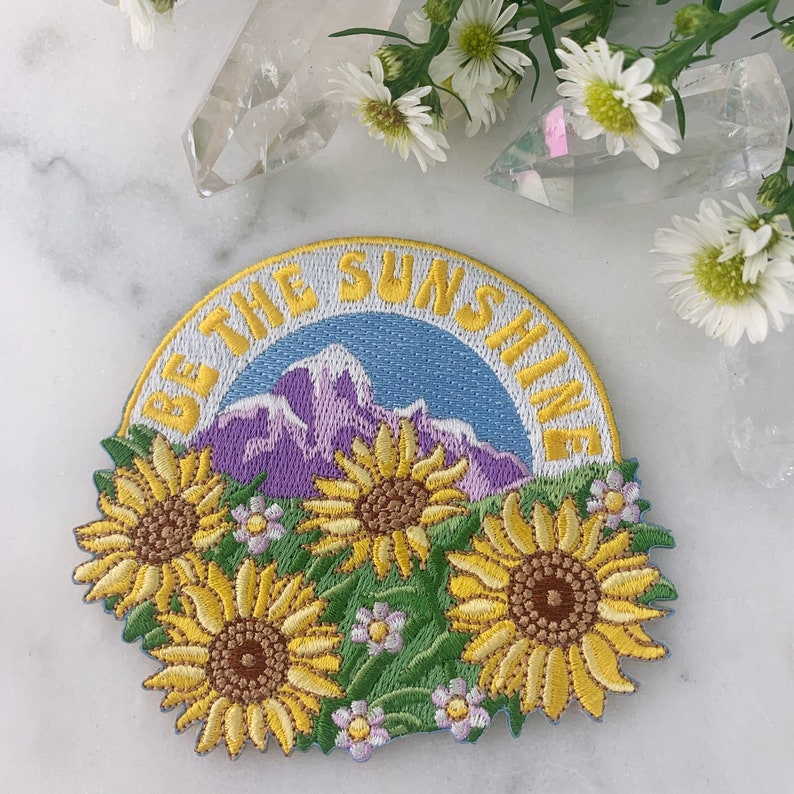 May include: Embroidered patch with a yellow, blue, and purple mountain scene and the text "Be the Sunshine". The patch is surrounded by yellow sunflowers and white flowers.