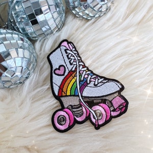May include: Embroidered patch of a white roller skate with a rainbow and pink heart design. The skate has pink wheels and white laces. The patch is set against a fluffy white background with disco ball ornaments.