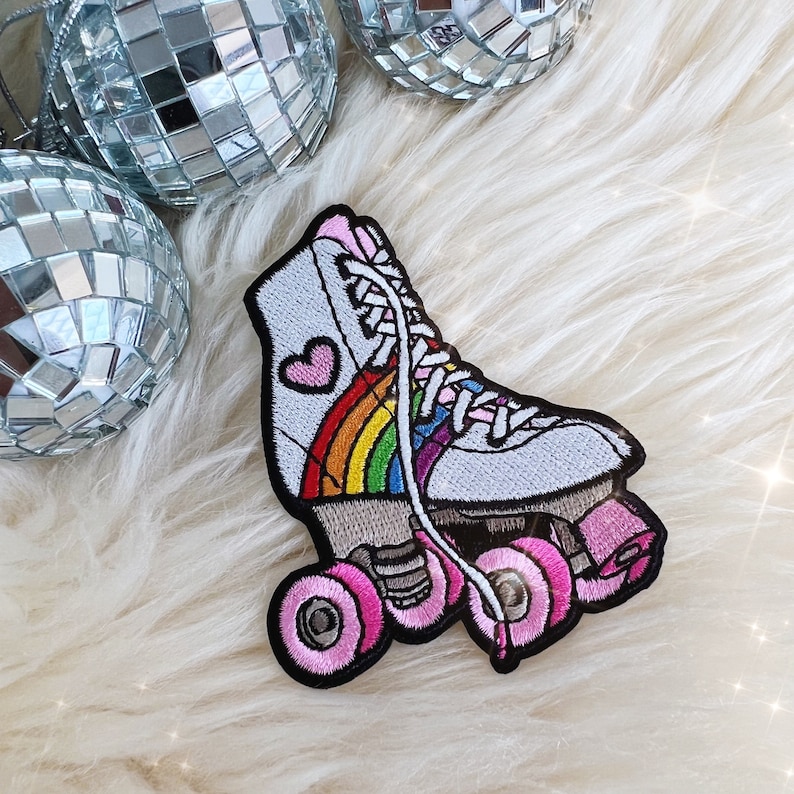 May include: Embroidered patch of a white roller skate with a rainbow and pink heart design. The skate has pink wheels and white laces. The patch is set against a fluffy white background with disco ball ornaments.