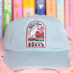 May include: Pale blue baseball cap with an embroidered patch. The patch shows a white cat reading a book on a stack of books. Text on the patch reads "There's no such thing as too many books."