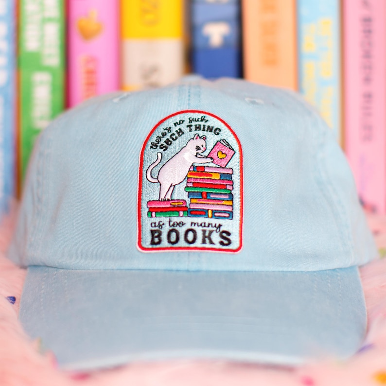 May include: Pale blue baseball cap with an embroidered patch. The patch shows a white cat reading a book on a stack of books. Text on the patch reads "There's no such thing as too many books."
