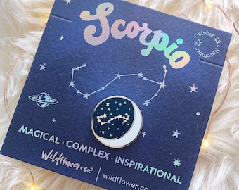 May include: A black and gold enamel pin featuring the constellation Scorpio with a crescent moon. The pin is on a blue background with white stars and the text "Scorpio" in a holographic font. The text "October 23 - November 22" is also on the background. The text "MAGICAL. COMPLEX. INSPIRATIONAL" is at the bottom of the background. The text "Wildflower+co" and "wildflower.co" are also at the bottom of the background.