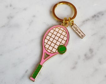 May include: A pink and gold tennis racket keyring with a green tennis ball. The keyring has a gold ring and a small tag that says "Sweet/Flower + Co."