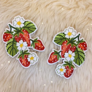 Könnte beinhalten: Zwei gestickte Bügelbilder mit Erdbeeren und weißen Blüten. Die Patches befinden sich auf einem weißen, flauschigen Hintergrund.