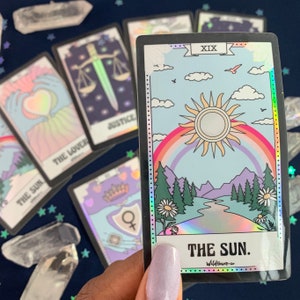 Puede incluir: Una carta de tarot hologr&aacute;fica con un borde negro y un dise&ntilde;o de arco&iacute;ris. La carta representa un sol con un arco&iacute;ris sobre &eacute;l, un r&iacute;o que fluye a trav&eacute;s de un valle y monta&ntilde;as en el fondo. La carta est&aacute; etiquetada como "XIX" y "THE SUN."