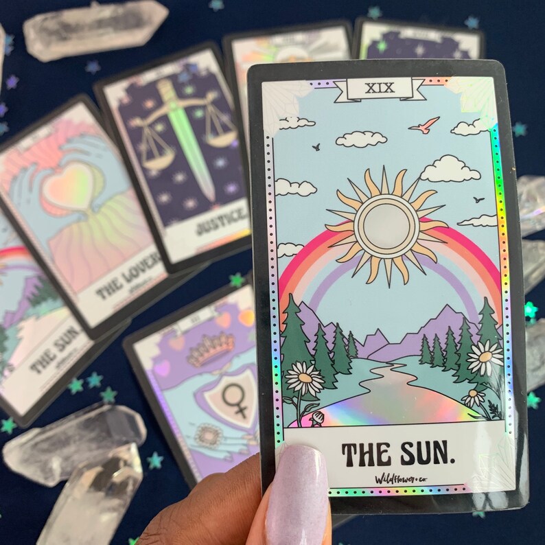 Puede incluir: Una carta de tarot hologr&aacute;fica con un borde negro y un dise&ntilde;o de arco&iacute;ris. La carta representa un sol con un arco&iacute;ris sobre &eacute;l, un r&iacute;o que fluye a trav&eacute;s de un valle y monta&ntilde;as en el fondo. La carta est&aacute; etiquetada como "XIX" y "THE SUN."