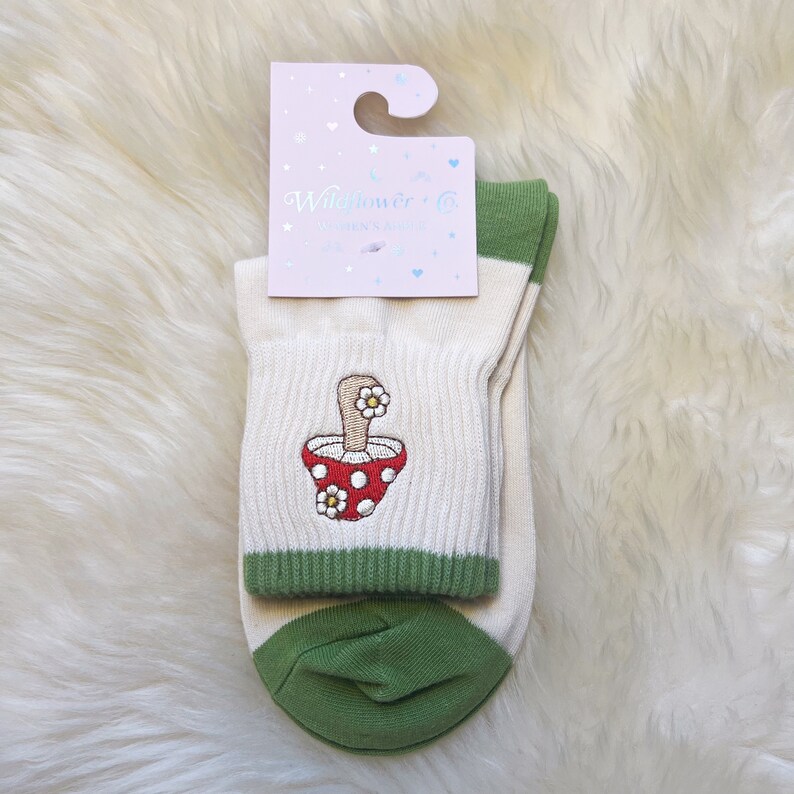 K&ouml;nnte beinhalten: Ein Paar wei&szlig;e Socken mit gr&uuml;nem Besatz und einem roten und wei&szlig;en Pilz mit einer wei&szlig;en Blume, die auf der Vorderseite gestickt ist. Die Socken sind mit einem rosa Etikett verpackt, auf dem "Wildflower Co. Clothing & Goods" steht.
