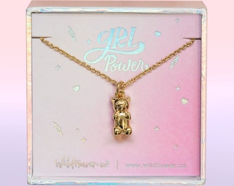 Puede incluir: Un collar con colgante de oso de peluche dorado, presentado en una caja con un fondo degradado rosa y blanco. La caja presenta el texto "GRL POWER" y el nombre de la marca "Wildflower co."