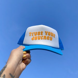 Könnte beinhalten: Eine blaue und weiße Trucker-Kappe mit weißem Netzrücken. Die Kappe trägt den Schriftzug "Trust Your Journey" in orangefarbenen Buchstaben auf der Vorderseite.