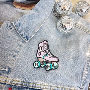 May include: A pink roller skate patch featuring a moon and star design, teal wheels, and white laces. The patch is on a light blue denim jacket, with three disco balls in the background.