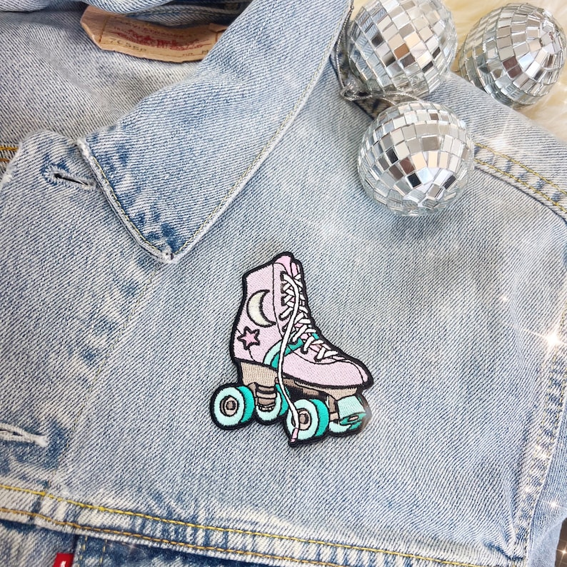May include: A pink roller skate patch featuring a moon and star design, teal wheels, and white laces. The patch is on a light blue denim jacket, with three disco balls in the background.