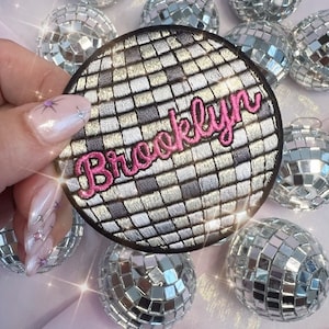May include: A round, silver and black embroidered patch with a mirrored disco ball design. The word "Brooklyn" is embroidered in pink cursive letters on the patch.