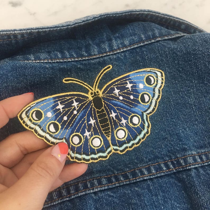 以下が含まれることがあります： 月と星のデザインが施された、青と金の刺繍の蝶のワッペン。ワッペンはアイロンで接着でき、衣服、バッグ、その他のアイテムの装飾に使用できます。