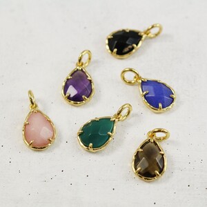 May include: Six gold teardrop-shaped pendants with different colored gemstones. The pendants are on a white surface. The gemstones are amethyst, black onyx, green agate, rose quartz, smoky quartz, and blue chalcedony.